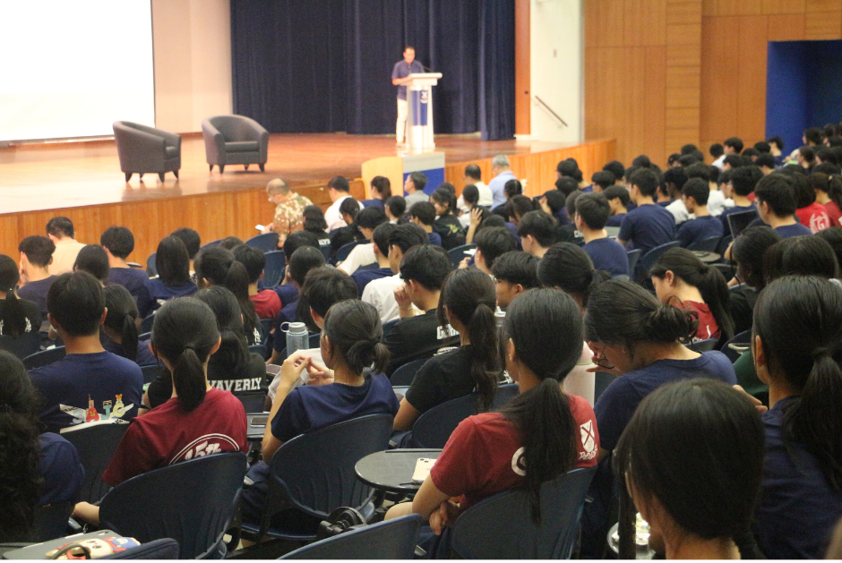 A large audience of students seated in an auditorium, viewed from behind, listening to SMS Mr Tan Kiat How at a podium on stage at St Andrew's Junior College.